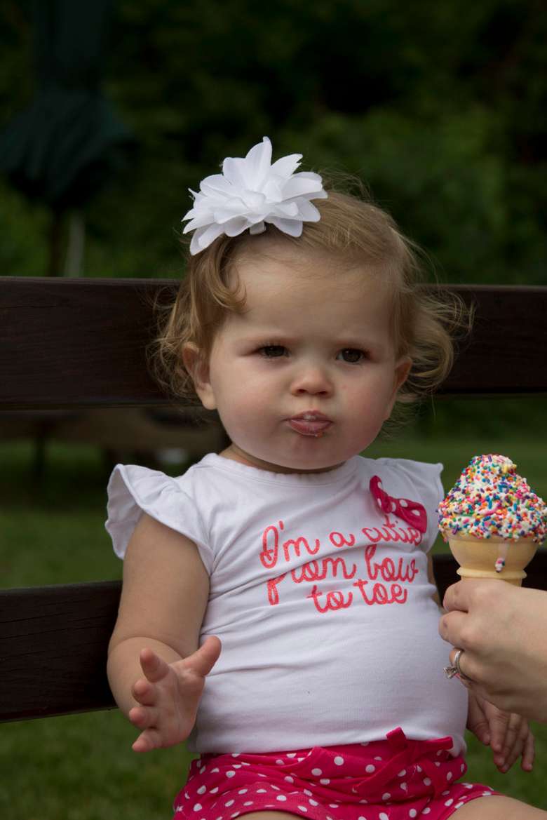 a toddler girl with a vanilla ice cream cone with sprinkles being handed to her, her shirt says I'm cute from bow to toe