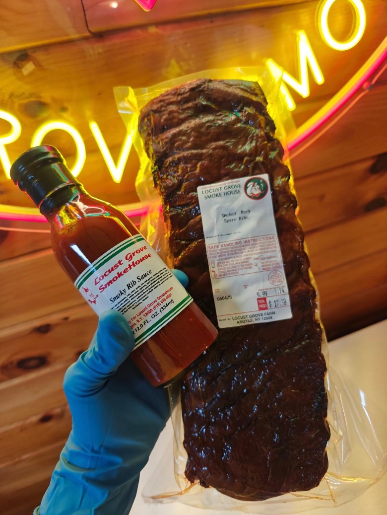 A gloved hand holding a bottle of Smoky Rib Sauce next to a vacuum-sealed rack of Smoked Pork Spare Ribs.