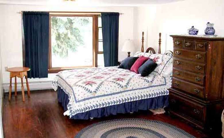 a bedroom with hardwood floors, bed with decorative quilt, large wooden chest of drawers and large window with blue drapes