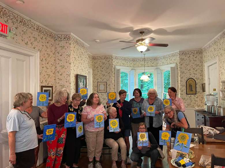 large group of women holding up paintings inside a house
