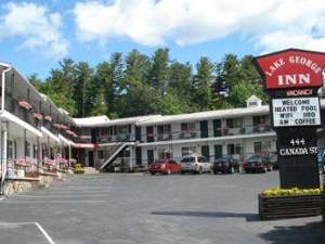 Outside view of the two-story motel. There are flowers in flower boxes along the railings on both levels. There is a sign that states Lake George Inn, Vacancy, Welcome, Heated Pool, WIFI, HBO, A.M. Coffee, 444 Canada Street.