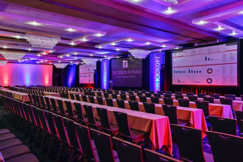 ballroom with long rectangular tables and two large projection screens set up for a conference or meeting