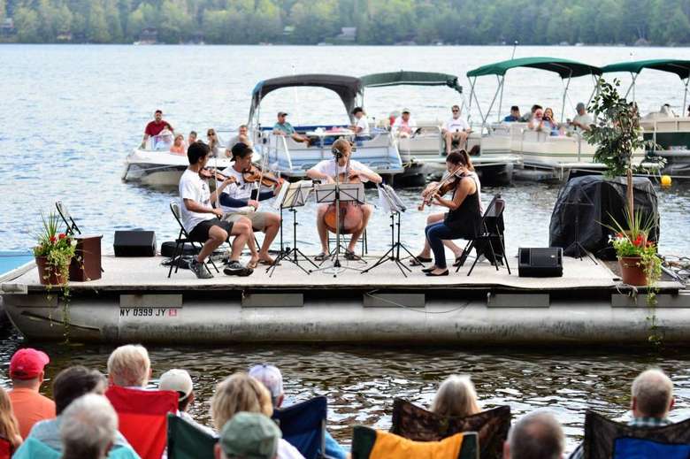 a four-piece group with string instruments perform on a boat in an open setting with other people on boats in the water watching them