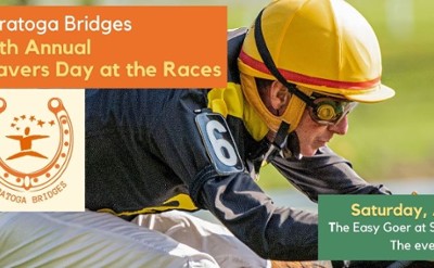 A jockey wearing a yellow hat riding a horse in a race with the Saratoga Bridges logo surrounded by a horseshoe.