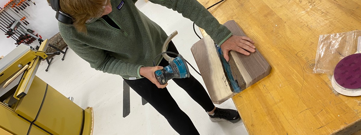 a woman sands her charcuterie board with a handheld orbital sander