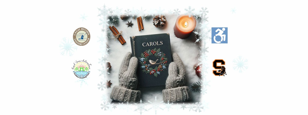 Two mittened hands are holding a book that says "Carols." Around the hands are a few cinnamon sticks, an anise star, and a candle.