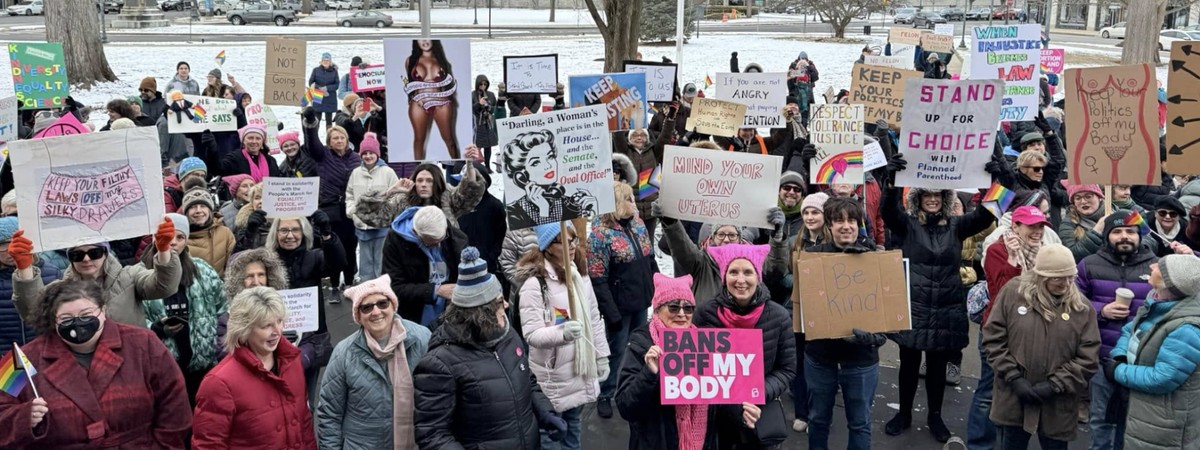 A large crowd gathers outdoors in winter, holding signs supporting women's rights and reproductive freedom. Many wear pink hats and hold rainbow flags. Snow-covered ground and trees in the background.