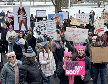 A large crowd gathers outdoors in winter, holding signs supporting women's rights and reproductive freedom. Many wear pink hats and hold rainbow flags. Snow-covered ground and trees in the background.