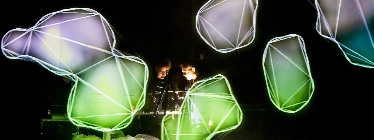 Two musicians play a keyboard, surrounded by colorful, light-up rock shapes that illuminate the scene.