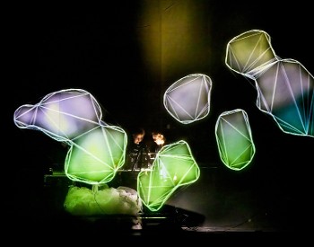 Two musicians play a keyboard, surrounded by colorful, light-up rock shapes that illuminate the scene.
