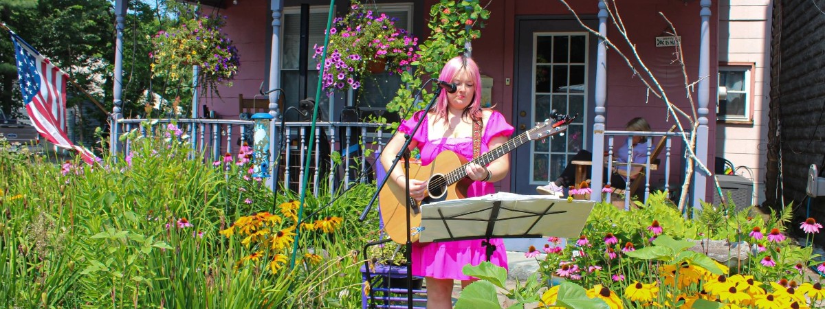 Woman in pink dress with pink hair stands in garden in front of pink house playing guitar with a microphone and music stand