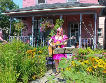 Woman in pink dress with pink hair stands in garden in front of pink house playing guitar with a microphone and music stand