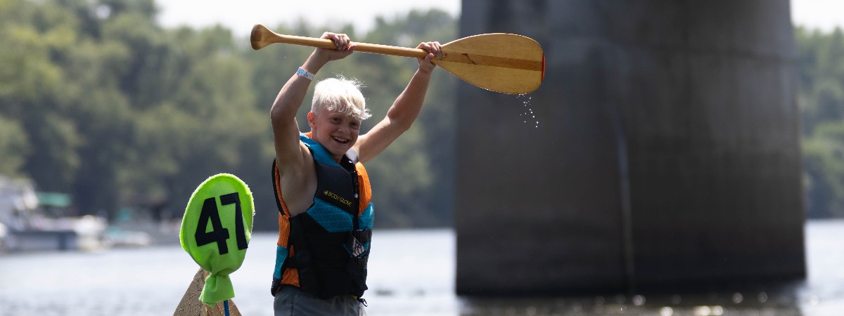 A boy holds his oar over his head in victory as he comes across the finish line during the 2025 Cardboard Boat Races.