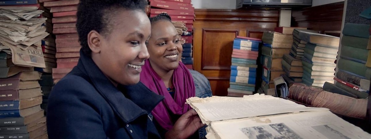 Two Nairobi women smiling as one lady holds an open book, there are piles of books and papers in the background.
