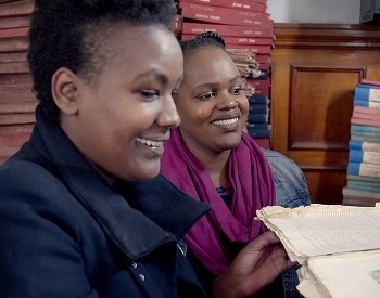 Two Nairobi women smiling as one lady holds an open book, there are piles of books and papers in the background.