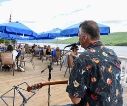 man playing guitar in front of a crowd of people sitting at tables next to the lake