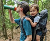 dad drinks from water bottlel, toddler in backpack carrier smiles