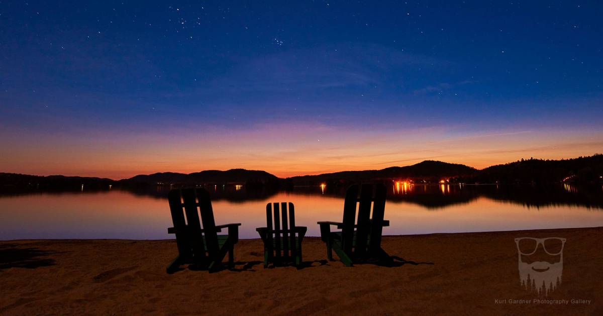 three Adirondack chairs in front of lake at sunset