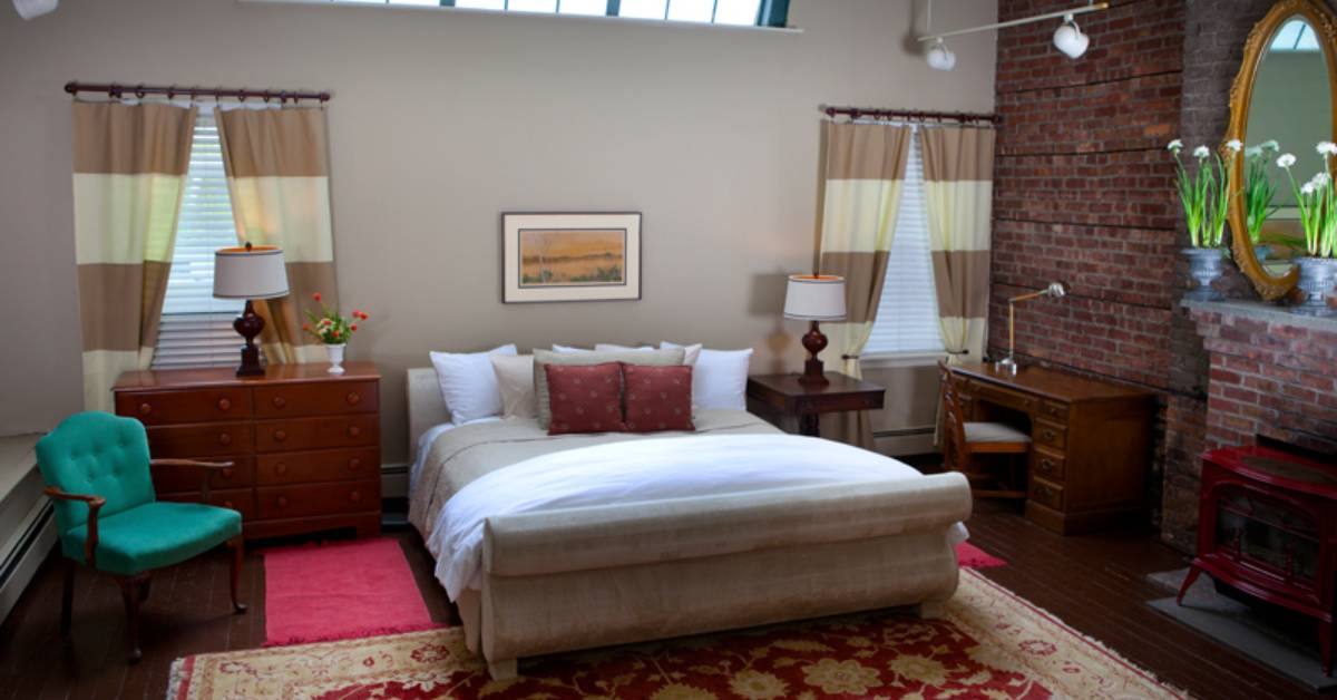 a guest room with a large bed, a green chair, a nightstand, dresser, and a red fireplace