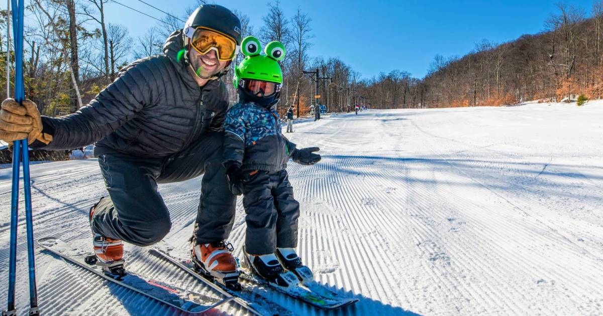 a dad and kid on skis, kid has green frog helmet