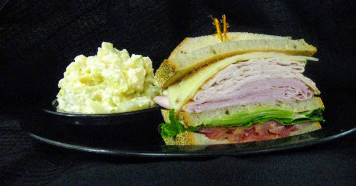 a stacked turkey, ham, cheese, lettuce, and tomato sandwich on a plate with a side of egg salad