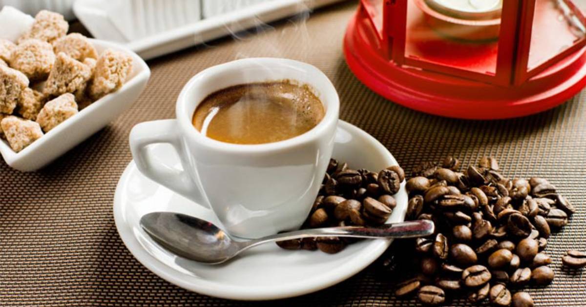 a cup of coffee with coffee beans spillin goff the saucer, what looks like scones to the left, and a small white candle in a red container on the right