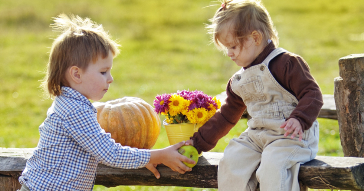 two little kids who look like brother and sister, the girl is sitting on a bench and the boy is handing her an apple