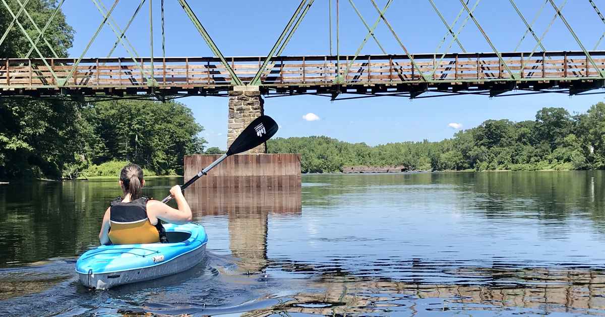 woman paddling in a blue kayak under a bridge part of a river