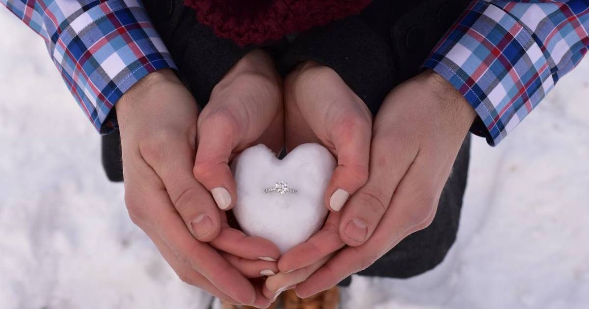 a couple's hands coming together to hold a heart-shaped piece of snow that has an engagement ring in it