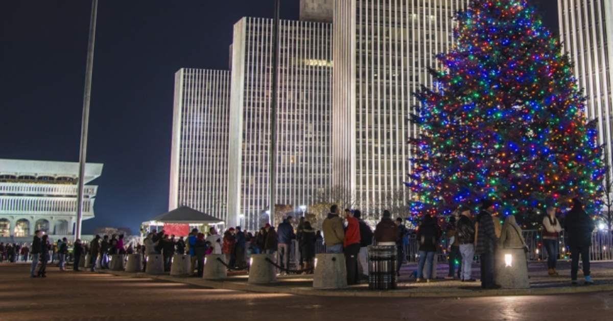 crowd standing outside city buildings near a large Christmas tree with lights