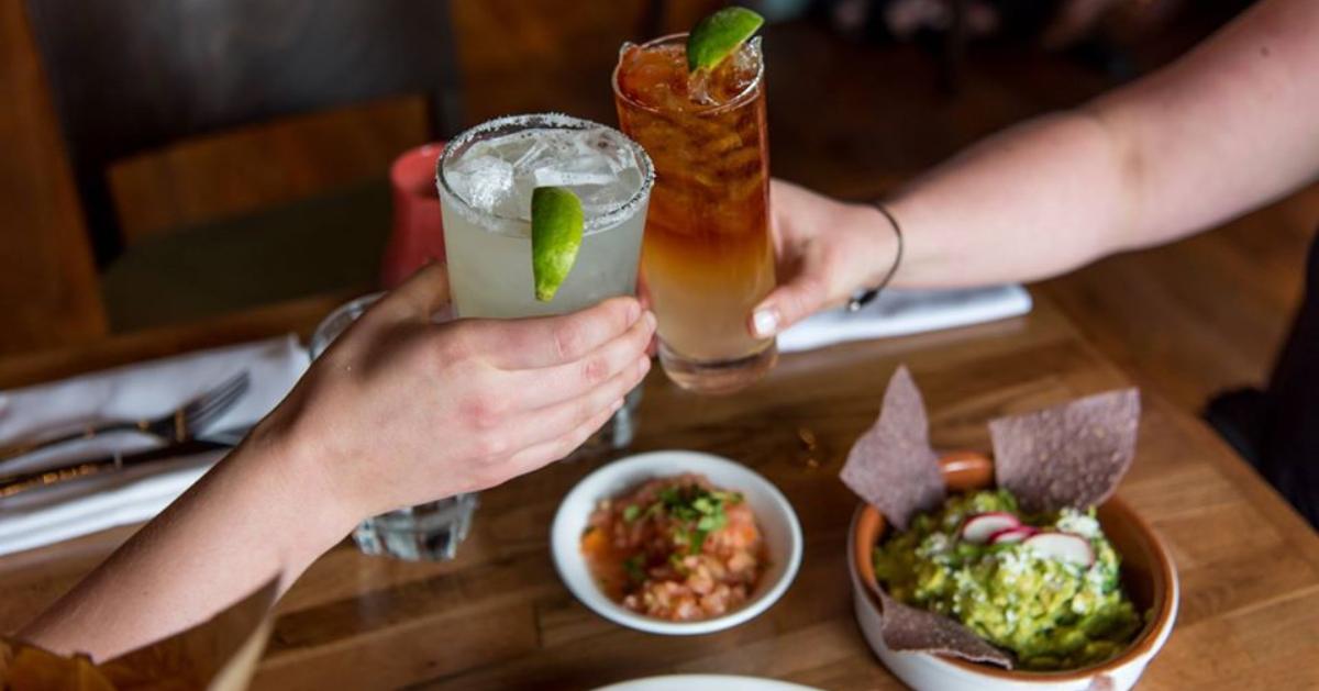 two people raising drinks over bowl of guacamole and chips