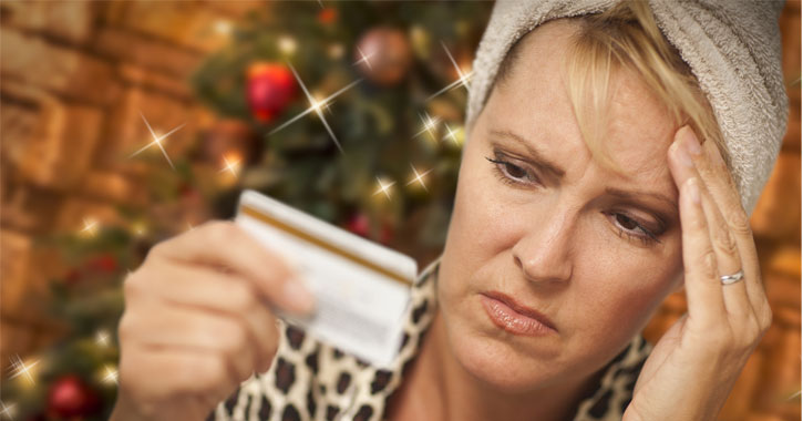 a woman looking pained holding up a credit card in front of a Christmas tree
