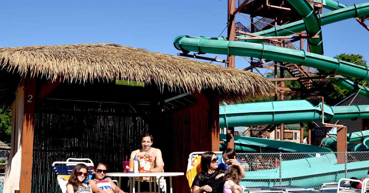 a family in a cabana at enchanted forest water safari, with a large waterslide in the background