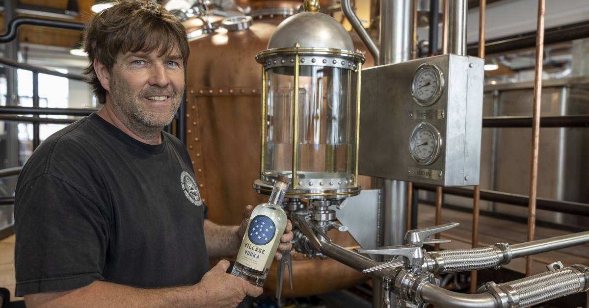 man holding up a bottle of village vodka by a copper distilling machine