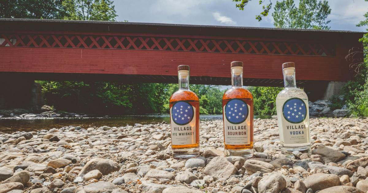 bottles of village rye whiskey, village bourbon, and village vodka on the ground below a red covered bridge