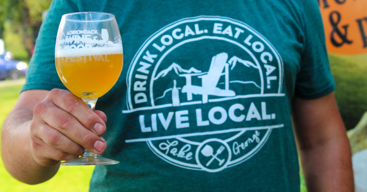 closeup of man holding adirondack wine and food festival glass, wearing drink local eat local live local shirt
