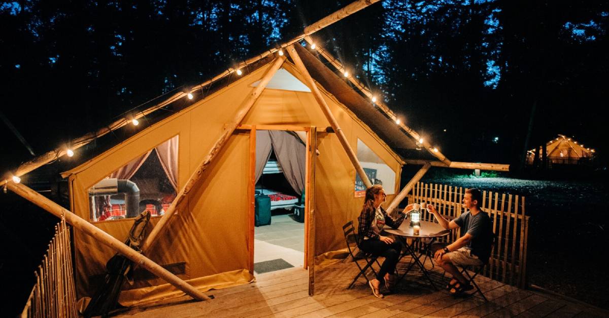 couple drinking beverages on a deck outside of a lit up glamping tent at night