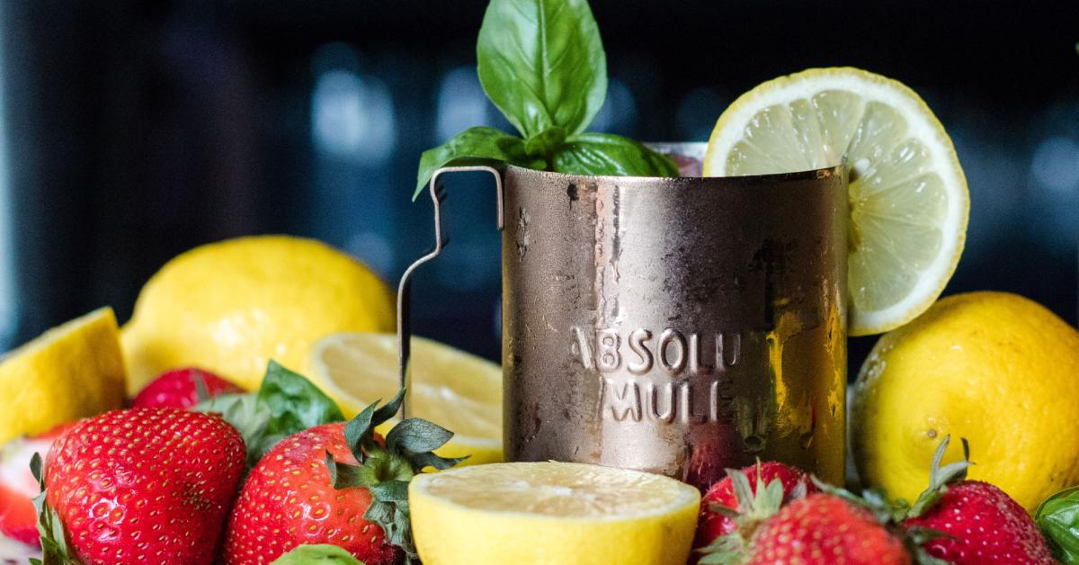 a drink in a mule mug with a mint sprig and lemon. lemons and strawberries around the glass