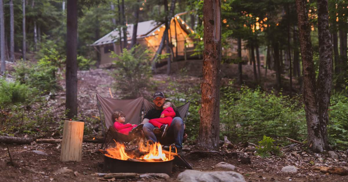 man and two kids sitting by a fire pit in the evening with a tent in the background