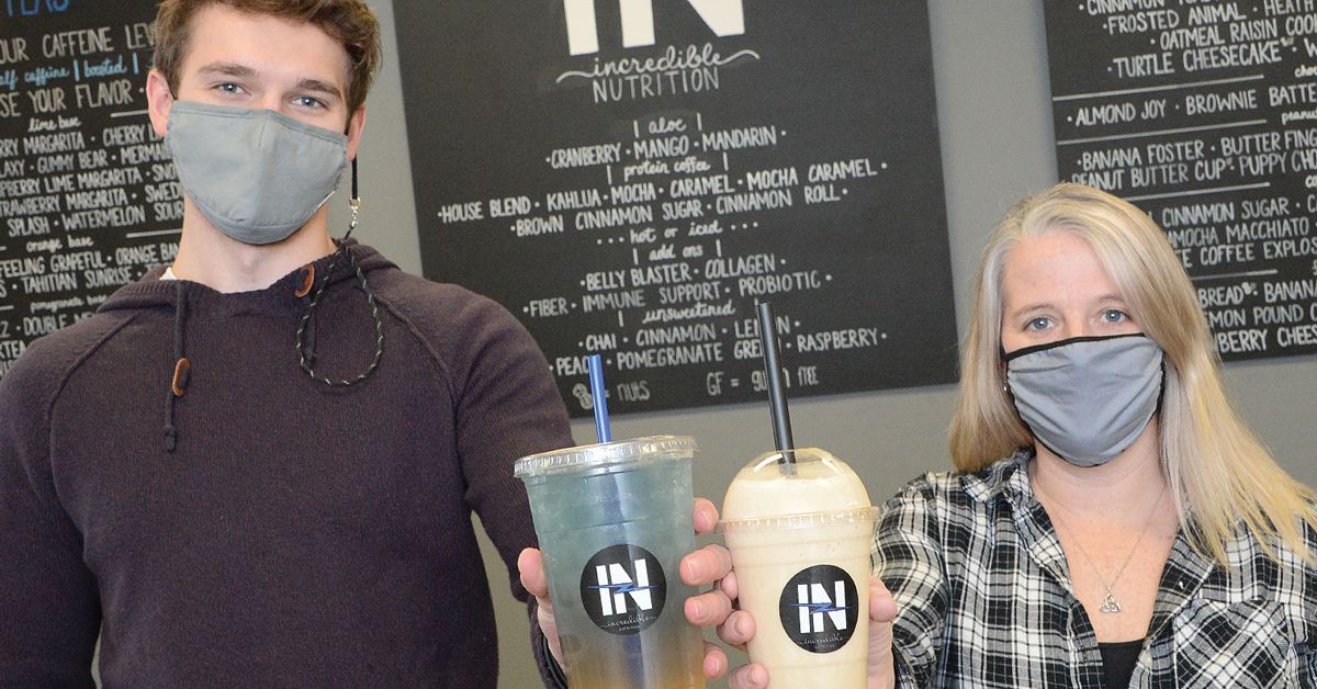 masked employees hold up iced tea and a shake, menus are behind them