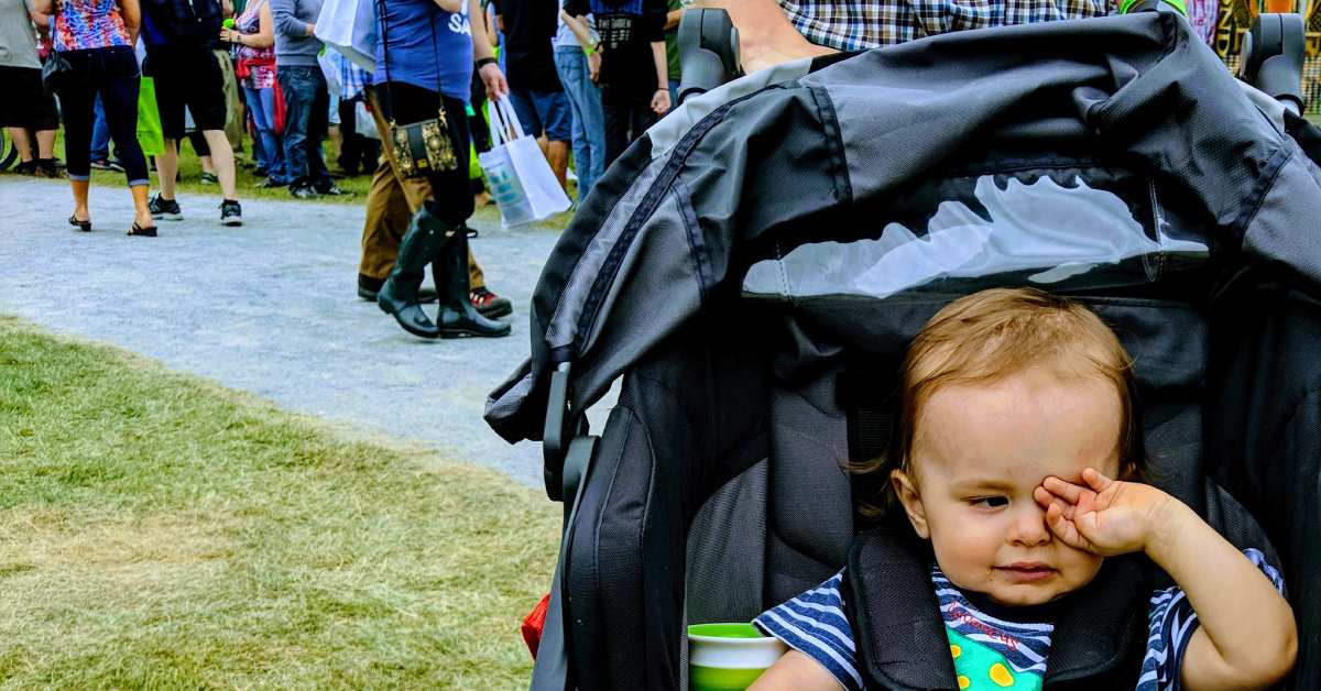 toddler in stroller in foreground rubs his eyes; people walking in background