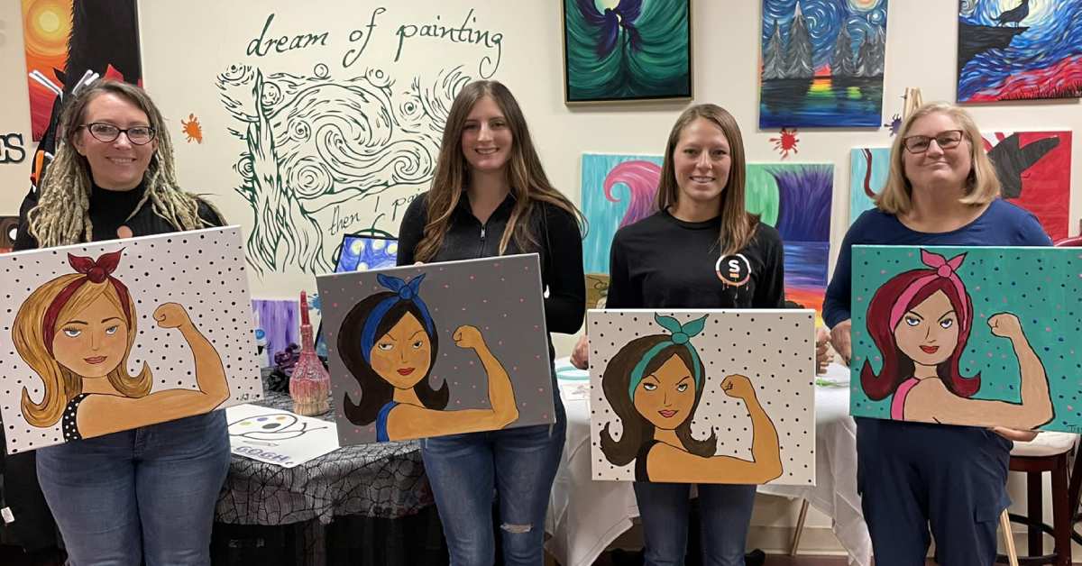 four women at paint and sip hold up paintings of women flexing an arm