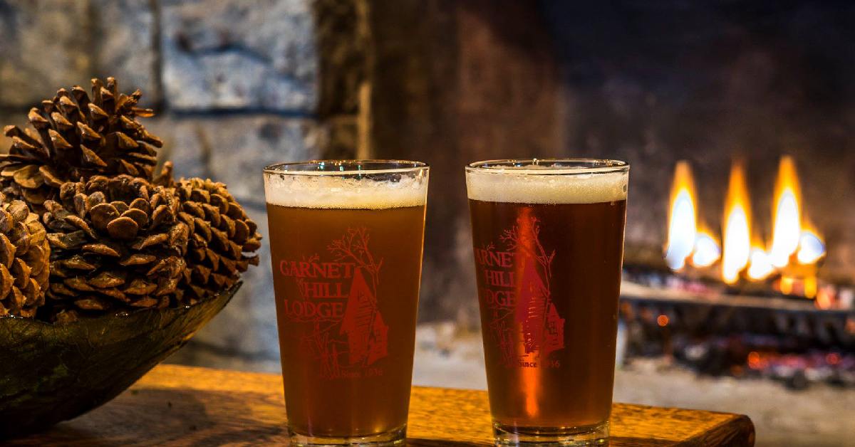 two beers and a basket of pinecones in front of a fireplace