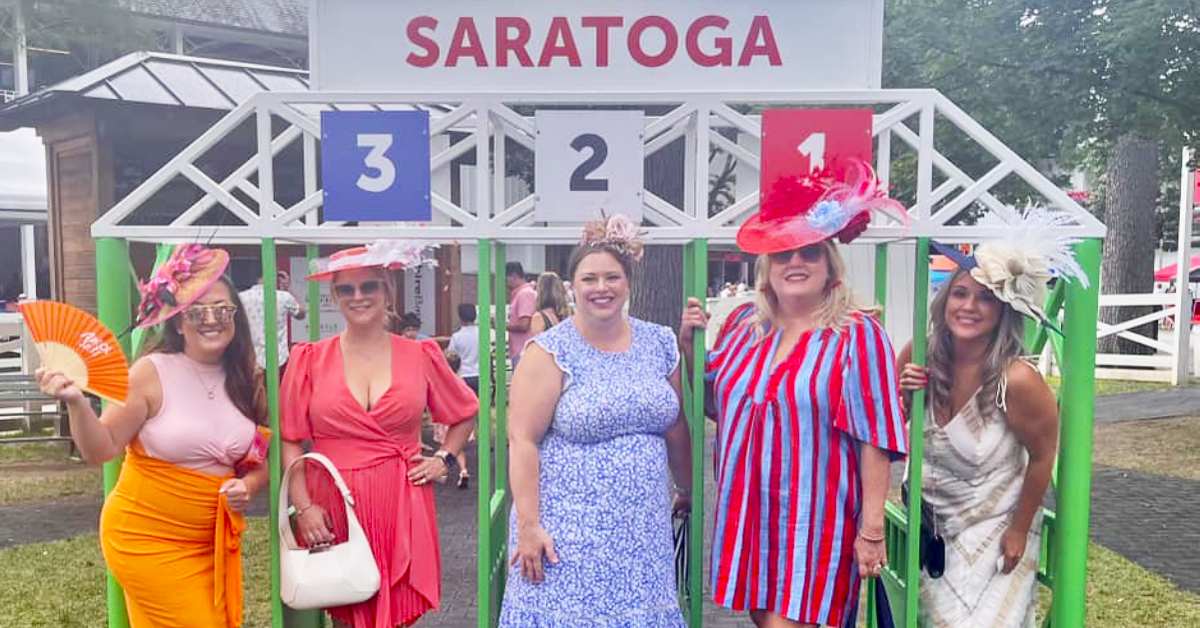 5 women dressed in bright colorful dresses and hats inside the mini horse starting gate