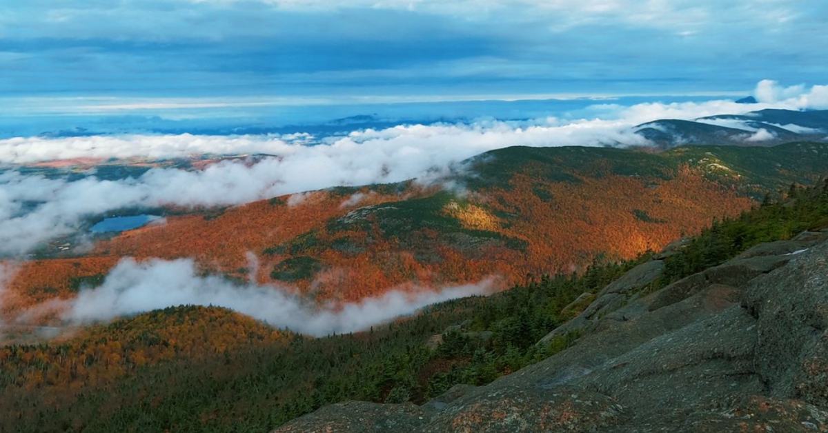 View of other mountains from a high peak that is above the clouds and looks over red, orange, and green trees