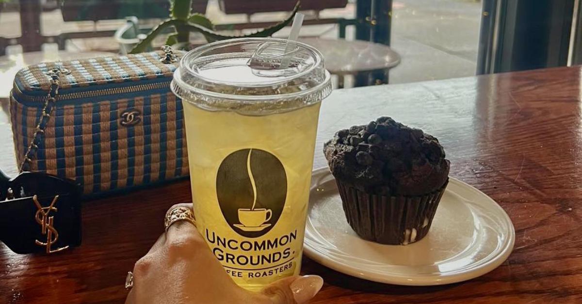 Hand holding ice tea on table next to a purse and chocolate muffin