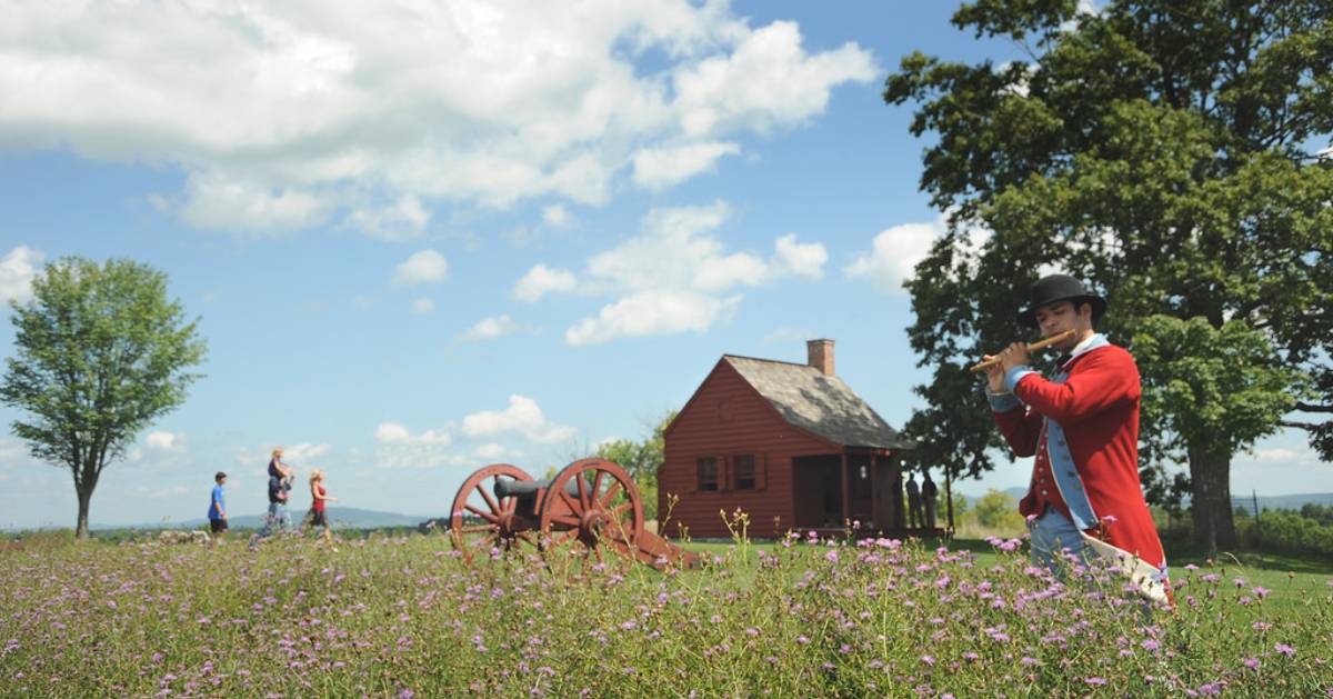 Field of flowers with man in red playing flute and a cannon and shack in the background
