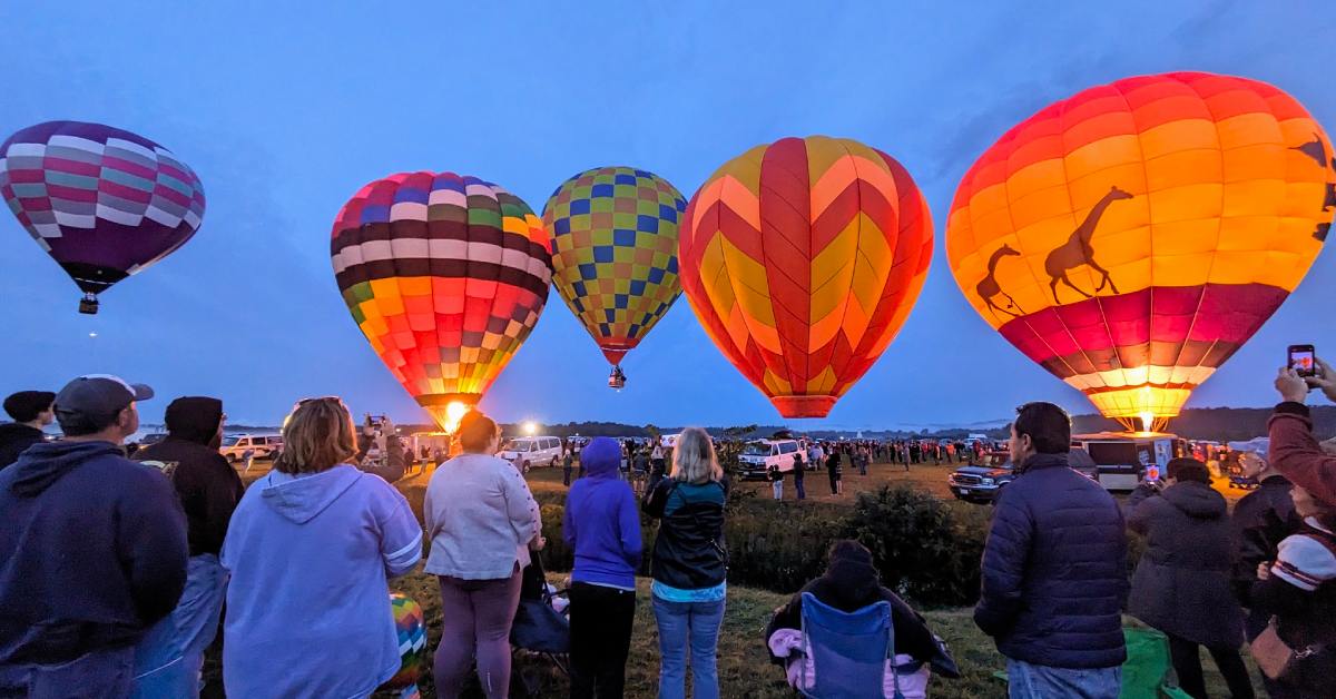 crowd watches as five hot air balloons start taking off into the night sky