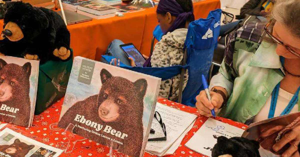 a woman signing a paper alongside a book called 'Ebony Bear'