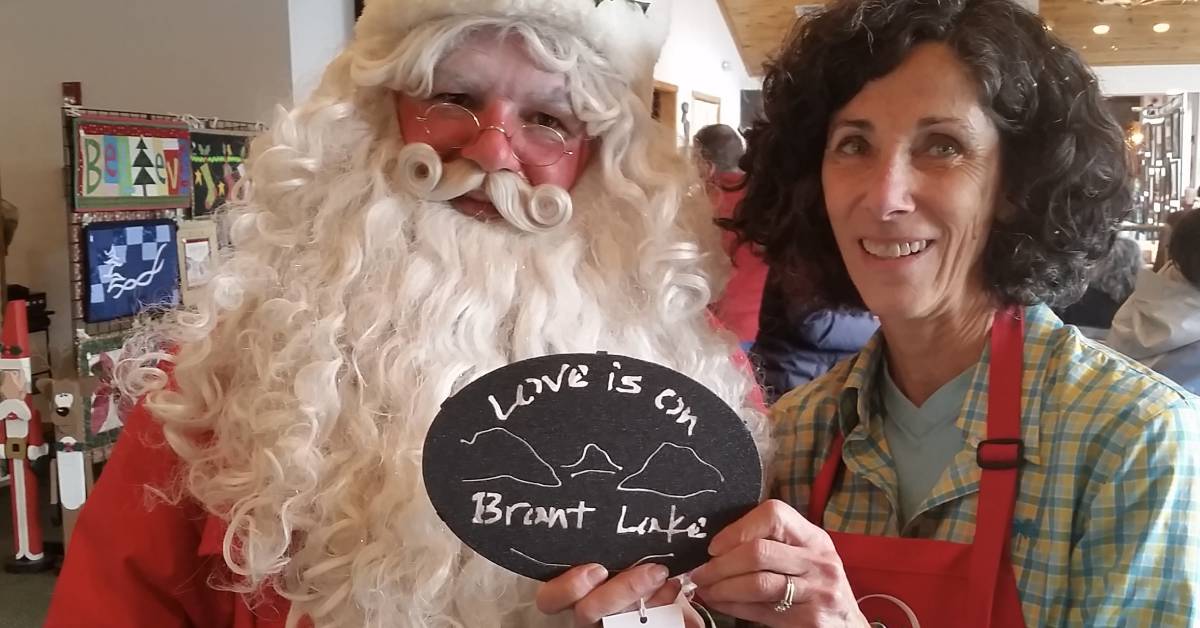 love is on lake george owner poses with santa while holding a love is on brant lake trivet